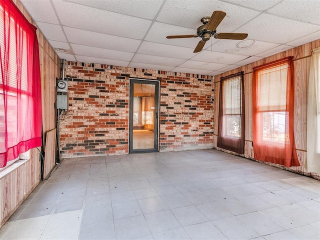 empty room with wooden walls, a drop ceiling, ceiling fan, and brick wall