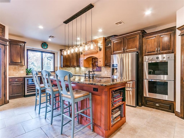 kitchen with a kitchen island with sink, stainless steel appliances, a kitchen breakfast bar, decorative light fixtures, and dark stone counters