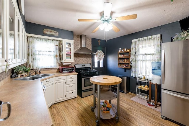 kitchen with a sink, white cabinetry, light wood-style floors, wall chimney range hood, and appliances with stainless steel finishes