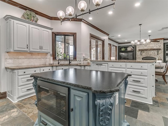 kitchen featuring stainless steel microwave, a stone fireplace, hanging light fixtures, ornamental molding, and a kitchen island