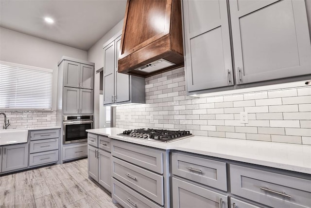 kitchen with sink, stainless steel appliances, gray cabinets, custom range hood, and light wood-type flooring