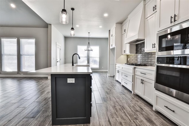 kitchen featuring custom exhaust hood, a kitchen island with sink, dark wood-type flooring, pendant lighting, and white cabinetry