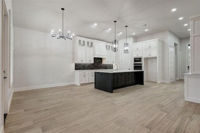 kitchen with decorative light fixtures, oven, a kitchen island with sink, built in microwave, and white cabinets