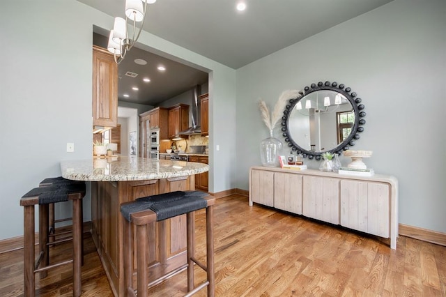 kitchen featuring kitchen peninsula, light stone countertops, light wood-type flooring, wall chimney exhaust hood, and hanging light fixtures