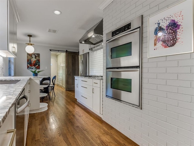 kitchen with light stone countertops, stainless steel appliances, wall chimney range hood, a barn door, and white cabinets