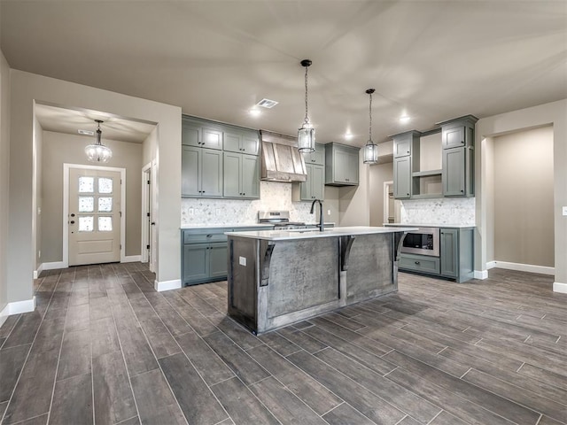 kitchen featuring custom exhaust hood, tasteful backsplash, a center island with sink, appliances with stainless steel finishes, and pendant lighting