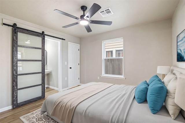 bedroom featuring light wood-type flooring, a barn door, ensuite bathroom, and ceiling fan