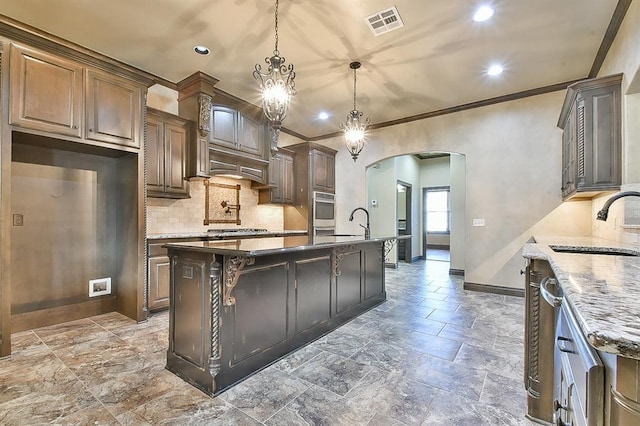 kitchen featuring a center island with sink, arched walkways, dark stone counters, pendant lighting, and a sink