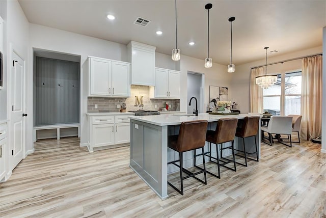 kitchen featuring decorative backsplash, light wood-type flooring, a kitchen island with sink, decorative light fixtures, and white cabinetry