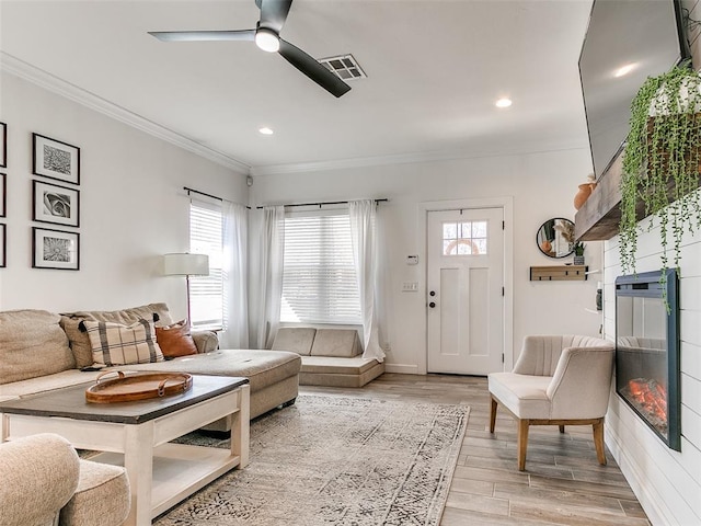 living area featuring recessed lighting, visible vents, light wood finished floors, a glass covered fireplace, and crown molding