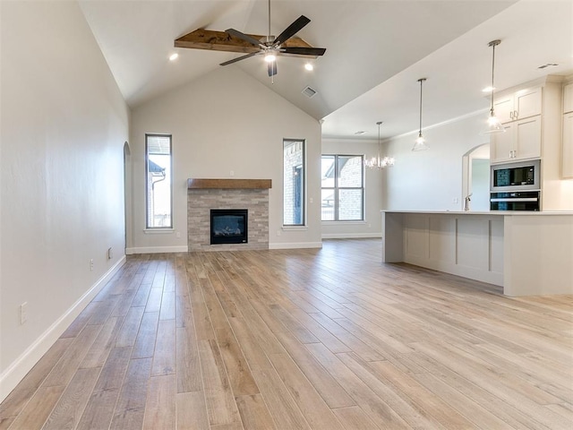 unfurnished living room featuring baseboards, light wood finished floors, high vaulted ceiling, a stone fireplace, and ceiling fan with notable chandelier