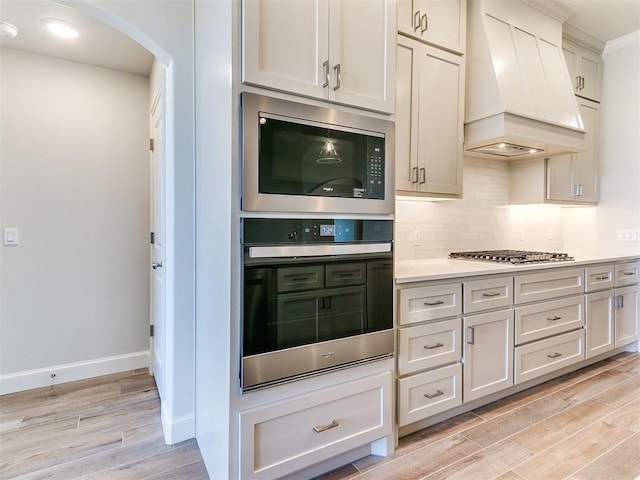 kitchen featuring light wood finished floors, backsplash, built in microwave, stainless steel gas cooktop, and custom range hood