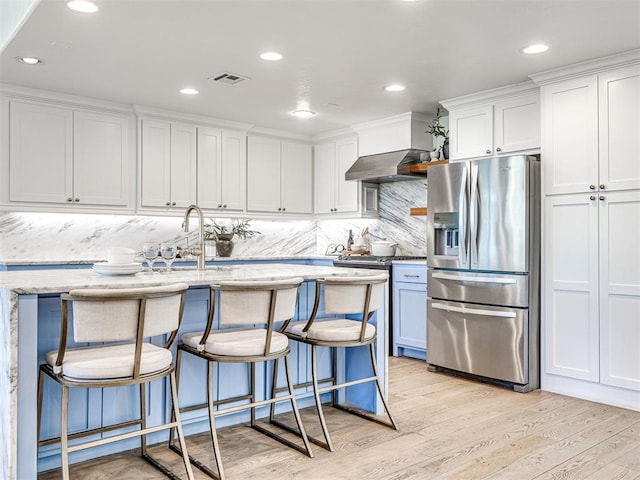 kitchen featuring white cabinetry, stainless steel fridge with ice dispenser, wall chimney range hood, and an island with sink