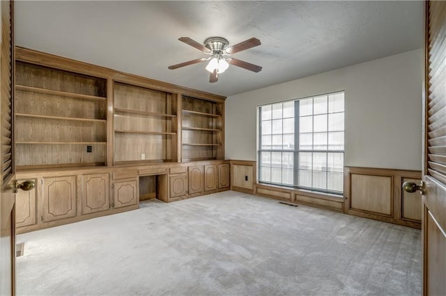 unfurnished living room with built in desk, light colored carpet, and ceiling fan