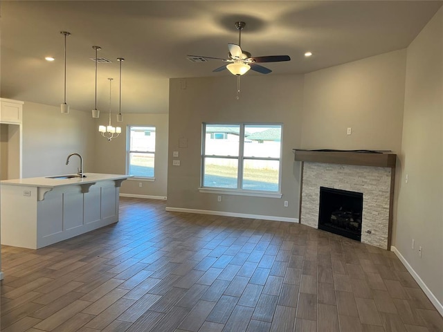 unfurnished living room with ceiling fan with notable chandelier, a stone fireplace, a sink, and dark wood finished floors