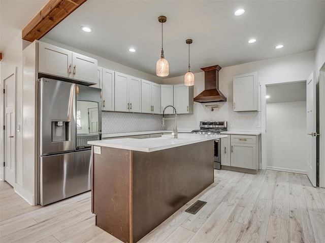 kitchen with stainless steel appliances, a kitchen island with sink, wall chimney range hood, light hardwood / wood-style flooring, and hanging light fixtures