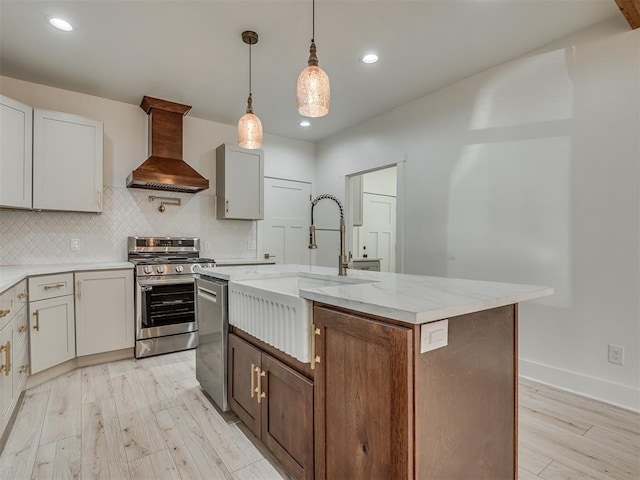 kitchen featuring appliances with stainless steel finishes, light hardwood / wood-style flooring, custom range hood, and a kitchen island with sink