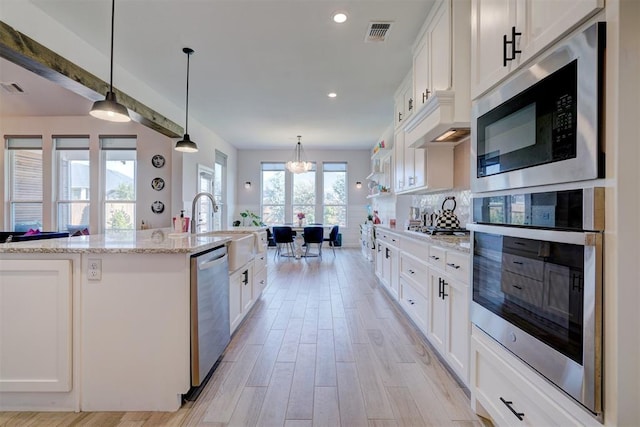 kitchen with appliances with stainless steel finishes, decorative light fixtures, white cabinets, light hardwood / wood-style floors, and an island with sink