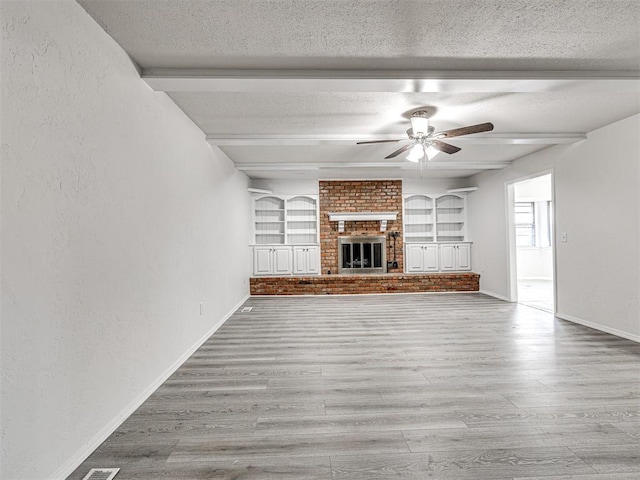 unfurnished living room with a brick fireplace, ceiling fan, a textured ceiling, beamed ceiling, and light hardwood / wood-style floors