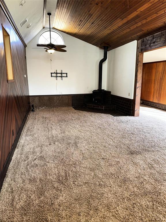 empty room with lofted ceiling, carpet floors, a wood stove, ceiling fan, and wooden ceiling