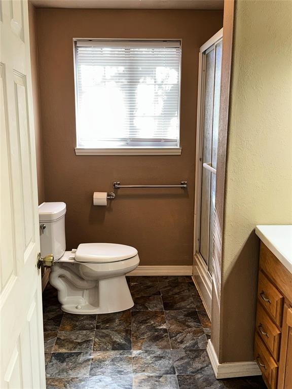 bathroom with a shower with door, toilet, stone finish floor, vanity, and baseboards