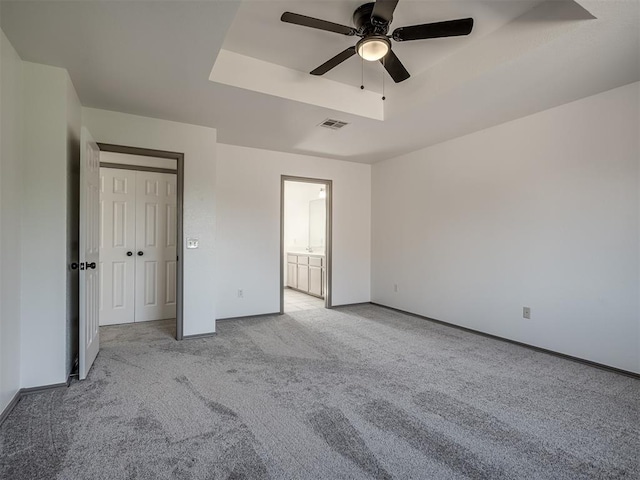 unfurnished bedroom with a raised ceiling, ceiling fan, ensuite bathroom, and light colored carpet