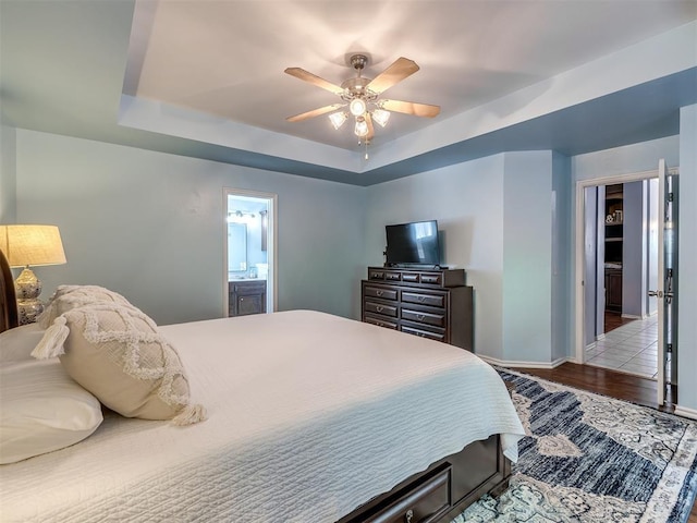 bedroom with ceiling fan, a tray ceiling, ensuite bath, and light wood-type flooring