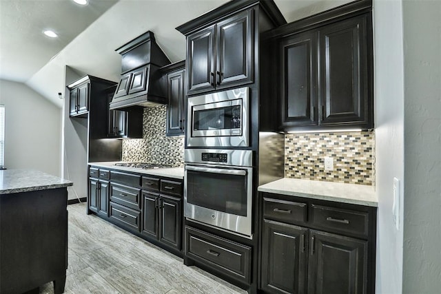 kitchen with appliances with stainless steel finishes, light wood-type flooring, backsplash, custom exhaust hood, and vaulted ceiling