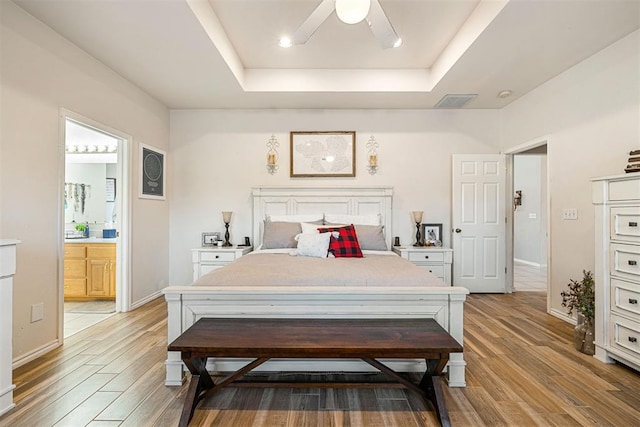 bedroom with ceiling fan, a raised ceiling, light wood-type flooring, and ensuite bathroom