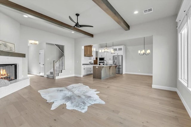 unfurnished living room with light hardwood / wood-style flooring, beamed ceiling, ceiling fan with notable chandelier, and a brick fireplace