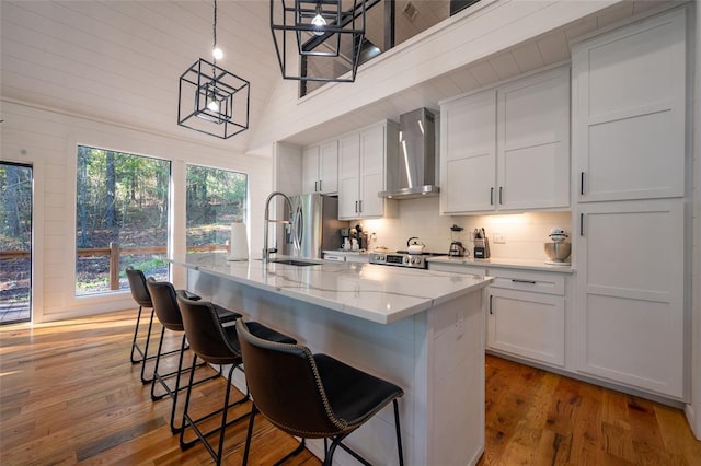 kitchen with a center island with sink, decorative light fixtures, wall chimney range hood, and high vaulted ceiling