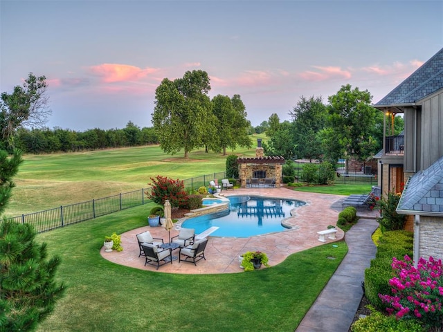 pool at dusk with an outdoor stone fireplace, a patio, and a lawn