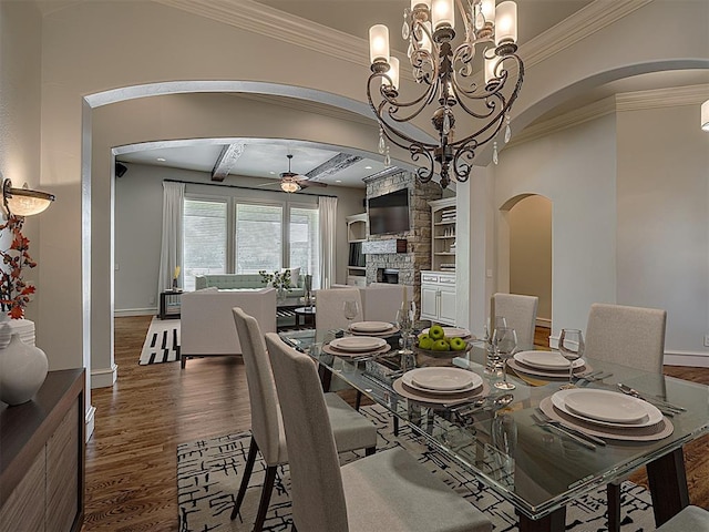 dining room with dark wood-type flooring, a stone fireplace, beamed ceiling, ceiling fan with notable chandelier, and ornamental molding