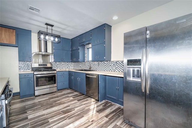 kitchen with blue cabinets, wall chimney range hood, sink, appliances with stainless steel finishes, and decorative light fixtures