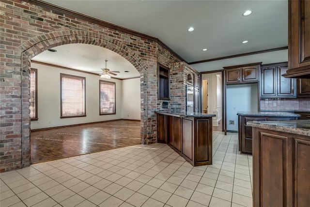 kitchen with dark stone counters, crown molding, ceiling fan, light tile patterned flooring, and brick wall