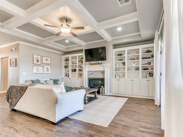 living room with light wood-type flooring, coffered ceiling, built in shelves, ceiling fan, and a stone fireplace