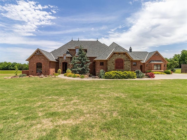 5451 Cottonwood Creek Cir, Norman, OK 73069 - french country home with a front yard, brick siding, a chimney, and roof with shingles