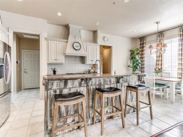 kitchen featuring stainless steel refrigerator with ice dispenser, backsplash, custom exhaust hood, pendant lighting, and white cabinetry