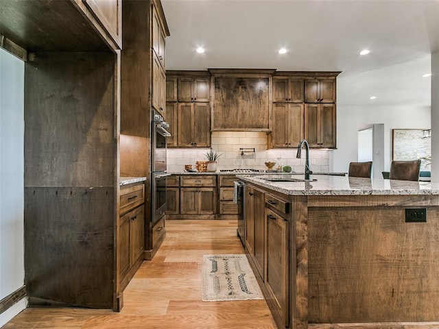 kitchen featuring light stone countertops, double oven, a kitchen island with sink, sink, and light hardwood / wood-style floors