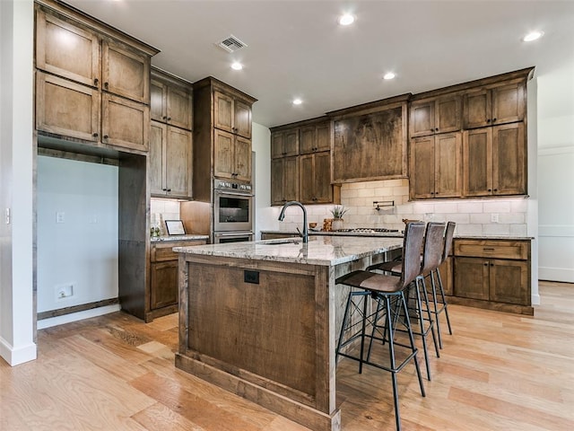 kitchen with light stone countertops, light wood-type flooring, sink, a center island with sink, and a breakfast bar area