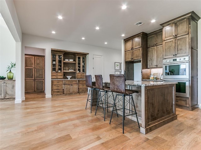 kitchen with light wood-type flooring, a center island with sink, a breakfast bar, light stone countertops, and double oven