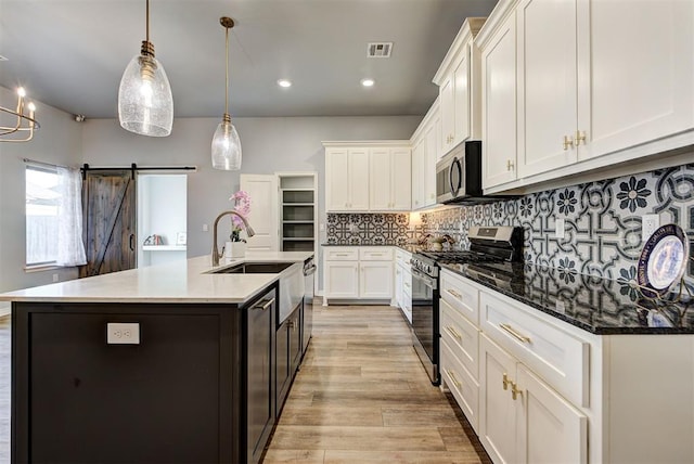 kitchen with appliances with stainless steel finishes, an island with sink, pendant lighting, a barn door, and white cabinets