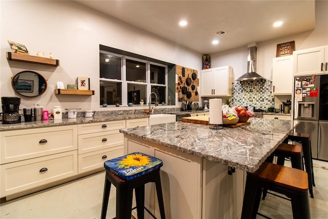 kitchen featuring a kitchen bar, stainless steel refrigerator with ice dispenser, light stone countertops, wall chimney exhaust hood, and a kitchen island