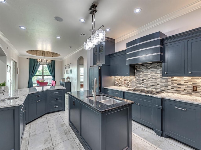 kitchen with stainless steel gas cooktop, premium range hood, hanging light fixtures, sink, and a center island with sink