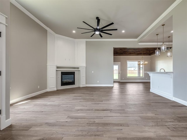 unfurnished living room with ceiling fan with notable chandelier, light wood finished floors, a glass covered fireplace, and crown molding