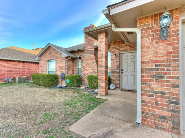 view of exterior entry featuring brick siding, a lawn, central AC unit, and a shingled roof