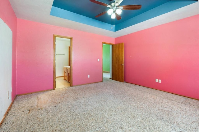 unfurnished bedroom featuring ensuite bath, ceiling fan, light colored carpet, a tray ceiling, and a walk in closet