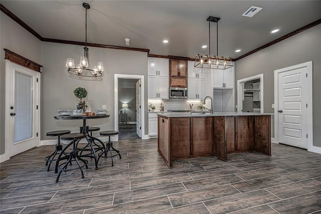 kitchen with decorative light fixtures, white cabinetry, a kitchen island with sink, and ornamental molding