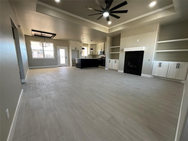 unfurnished living room with ornamental molding, baseboards, a raised ceiling, built in shelves, and ceiling fan with notable chandelier
