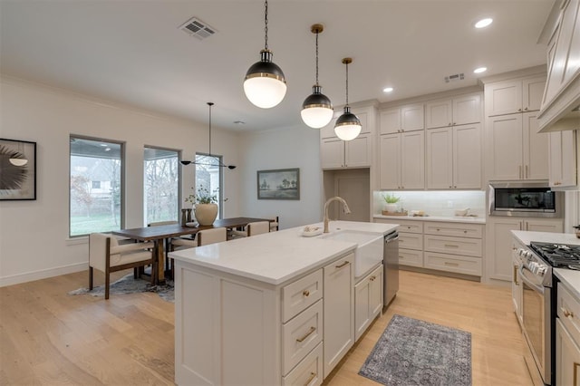 kitchen featuring light wood-type flooring, stainless steel appliances, a kitchen island with sink, sink, and hanging light fixtures